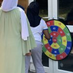 A girl throwing a ball at a colorful velcro target board attached to a glass door under adult supervision. - Copy (2)