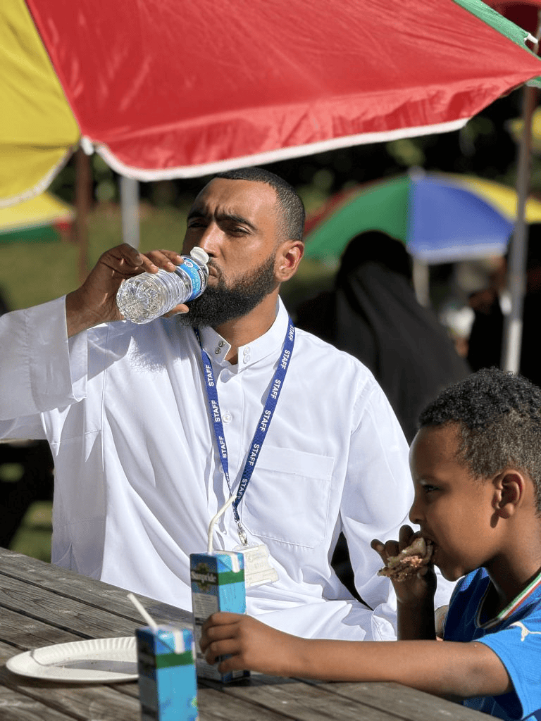 A man in a white thobe and staff lanyard drinks from a plastic water bottle at a wooden picnic table, while a young boy beside him eats a sandwich under colorful umbrellas outdoors.