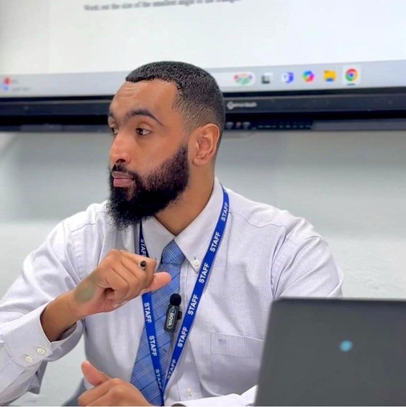 A man with a dark beard wearing a light blue dress shirt, patterned tie, and a blue lanyard labeled STAFF, seated in a classroom or office setting holding a marker in his hand