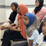 A row of young girls wearing headscarves sitting together in the audience, looking attentively forward_jfif