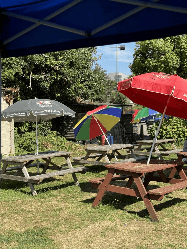 An outdoor patio area on a grassy lawn with several wooden picnic tables shaded by colorful sun umbrellas, including red, black, and multi-colored options.