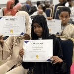 Children and young attendees hold up their printed certificates of achievement directly toward the camera during the indoor gathering_jfif