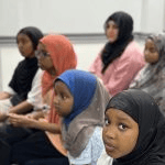 Displays young girls wearing headscarves seated together in the audience, looking forward attentively_jfif