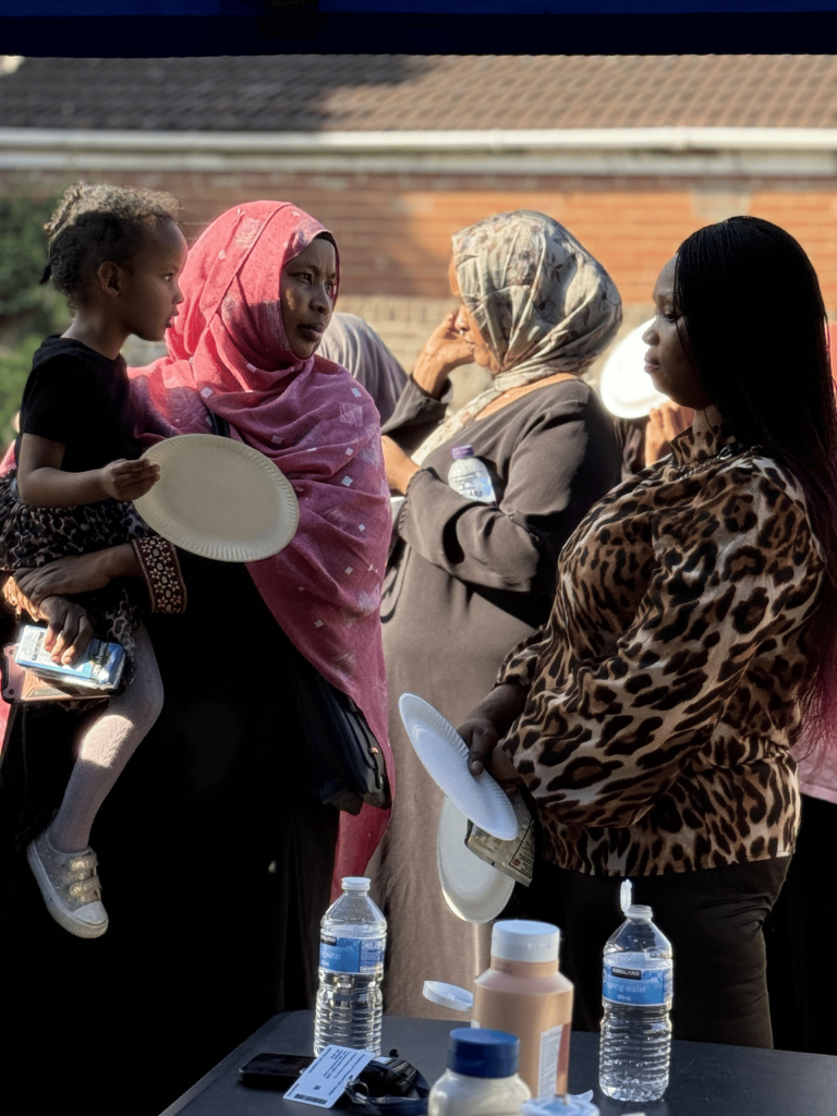 Group of women and a young child holding paper plates and conversing outdoors near a beverage setup.