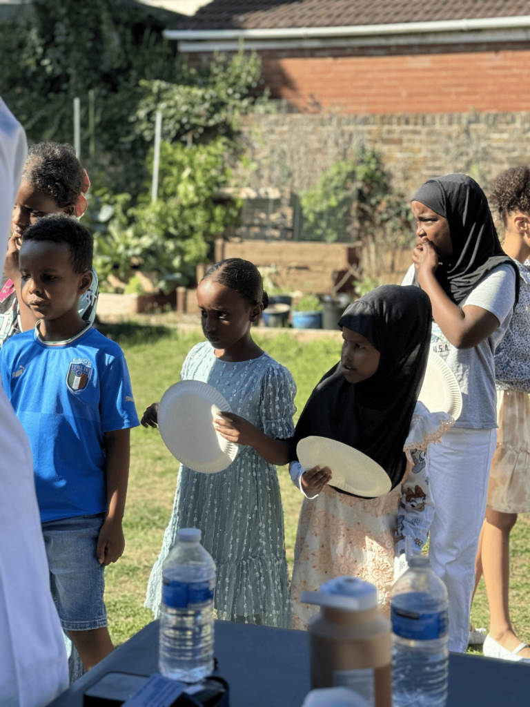 Group of young children standing outdoors in a queue holding empty paper plates, waiting to receive food at a sunny gathering.