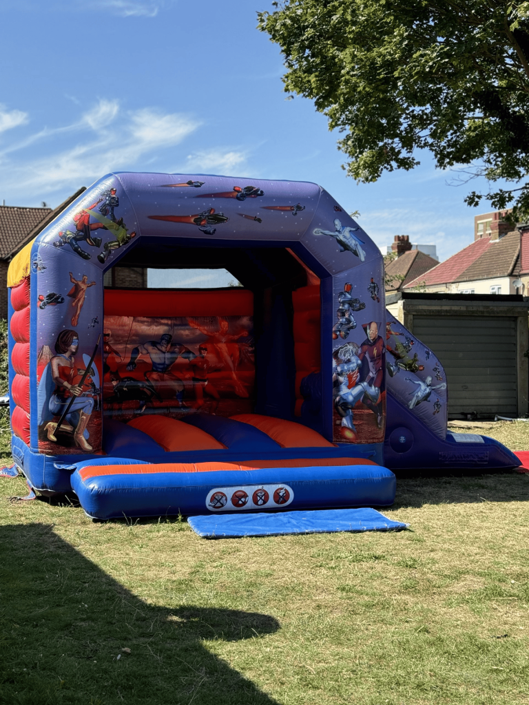 Inflatable blue and orange superhero-themed bouncy castle with an attached side slide set up outdoors on a grassy lawn under a sunny sky.