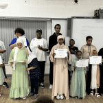 It shows a group of children and young adults standing together at the front of a roomhall holding their certificates of achievement_jfif