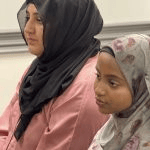 Two women wearing headscarves sitting attentively during an indoor gathering_jfif
