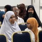 Women and children are seated in rows of chairs during an indoor ceremony, with a woman in the background capturing a photo on her smartphone_jfif